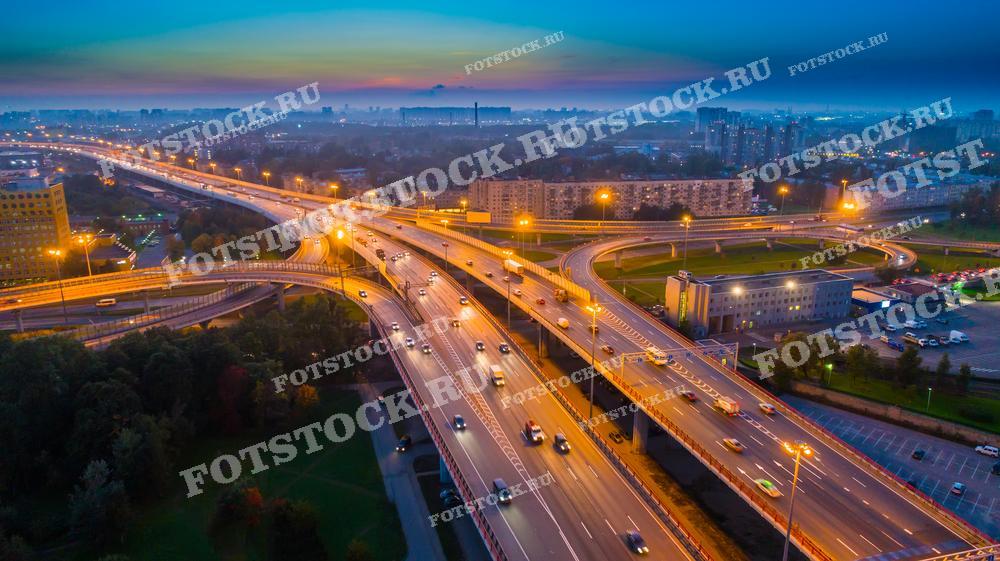Эта фотография демонстрирует утренний вид на оживлённую транспортную развязку в Санкт-Петербурге. Свет фонарей и утренние лучи создают динамичное освещение, подчёркивающее красоту урбанистического ландшафта. Фото идеально подходит для использования в материалах, связанных с градостроительством, транспортными исследованиями и городским планированием.